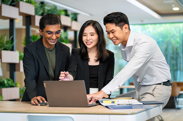 Three colleagues collaborating on a laptop, smiling in a modern workspace.